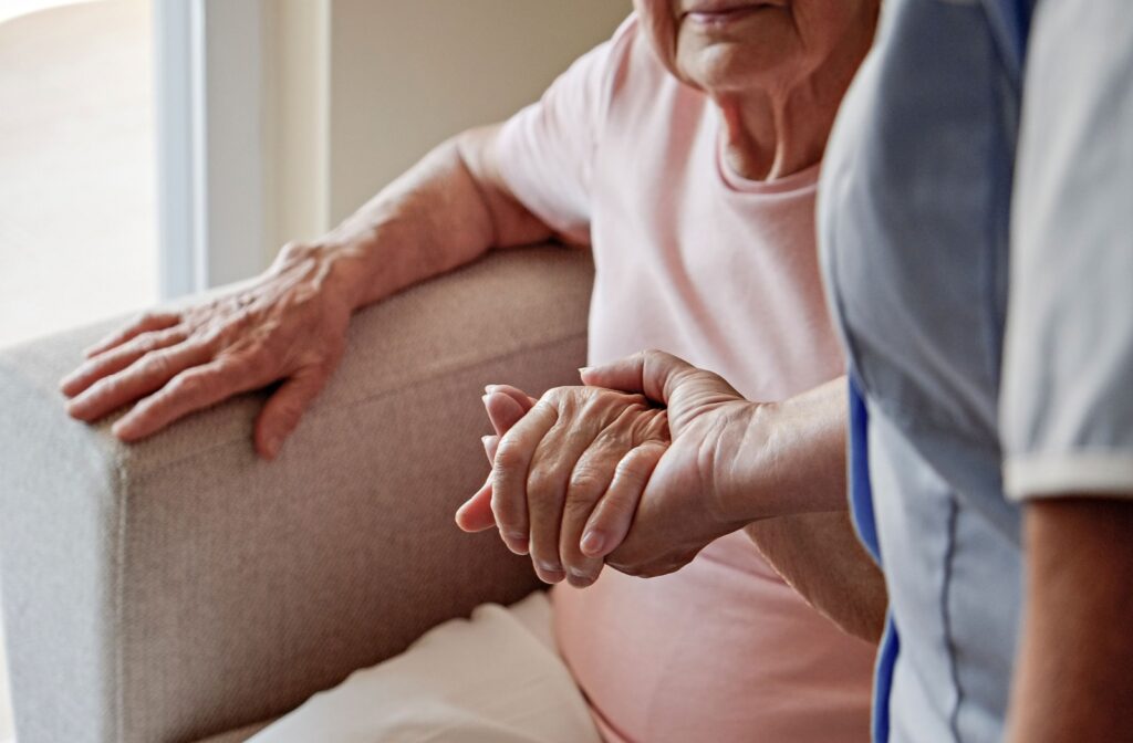 a close up of a caregiver holding the hand of an elderly woman as she helps her stand up from a couch.